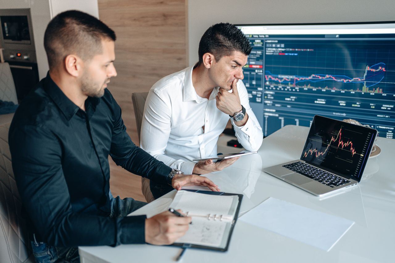 Two businessmen sitting at a desk analyzing stock charts on laptops and tablets.